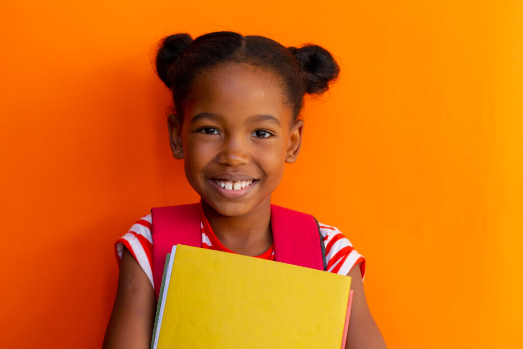 Our Curriculum portrait of happy african american schoolgirl with books over orange background at elementary school. education, childhood, development, learning and elementary school, unaltered.